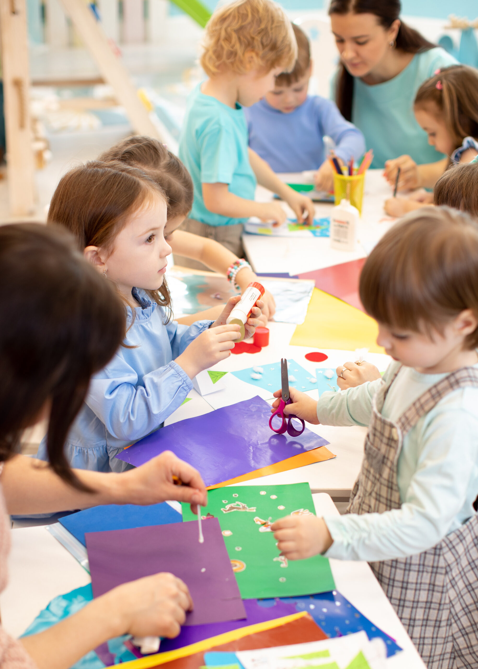 Development learning children in preschool. Children’s project in kindergarten. Group of kids and teacher cutting paper and gluing with glue stick on art class in kindergarten or daycare centre
